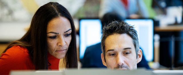 A woman and a man engaged in a focused discussion while working on a computer, symbolizing teamwork and strategic planning in social selling. The background features office monitors, representing a digital and data-driven work environment. | CBC | Cross-Border Communications | Leading international B2B branding and marketing agency